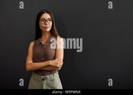 Business expert. Giovane donna asiatica in occhiali tenere le braccia incrociate e cercando in disparte mentre in piedi contro uno sfondo scuro. La gente di affari. Concetto di lavoro Foto Stock