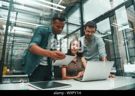 La ricerca di soluzioni. Un gruppo di tre giovani e positivo ai dipendenti che utilizzano le tecnologie moderne e discutere di qualcosa mentre si lavora in un ufficio moderno. Il concetto di lavoro. Luogo di lavoro Foto Stock