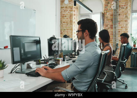 Focalizzati sul lavoro. Vista laterale di giovani occupati i dipendenti che lavorano su computer mentre è seduto alla scrivania in moderno spazio aperto. Il concetto di lavoro. Sul posto di lavoro. Lavoro di squadra Foto Stock