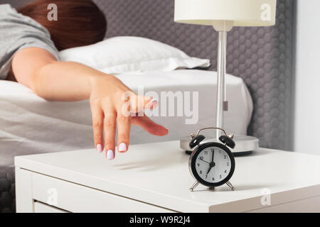 Donna di canto close-up cercando di raggiungere la Suoneria sveglia sul comodino la mattina presto. Al mattino il concetto di routine Foto Stock