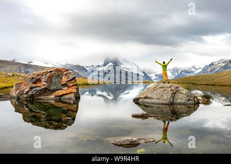 Escursionista sorge su rocce nel lago e si allunga le braccia in aria, Lago Stellisee, Cervino, nuvoloso, Vallese, Svizzera Foto Stock