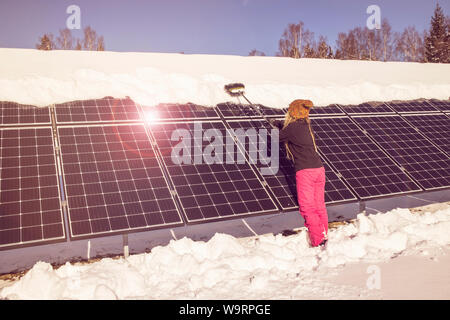 Donna spingendo la neve off pannelli solari in inverno. Se la neve copre i pannelli non sono in grado di produrre energia. Piccolo privato home casa sullo sfondo. Foto Stock