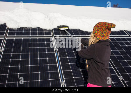 Donna spingendo la neve off pannelli solari in inverno. Se la neve copre i pannelli non sono in grado di produrre energia. Piccolo privato home casa sullo sfondo. Foto Stock