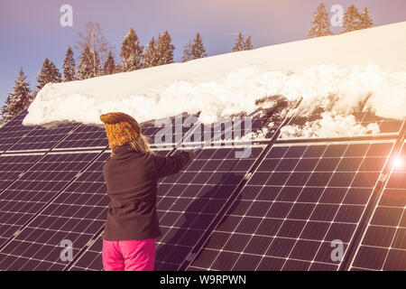 Donna spingendo la neve off pannelli solari in inverno. Se la neve copre i pannelli non sono in grado di produrre energia. Piccolo privato home casa sullo sfondo. Foto Stock