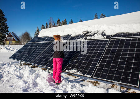 Donna spingendo la neve off pannelli solari in inverno. Se la neve copre i pannelli non sono in grado di produrre energia. Piccolo privato home casa sullo sfondo. Foto Stock