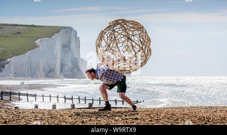 Cuckmere Haven, Regno Unito. 15 Agosto, 2019. Artista Keith Pettit pone il primo di una serie di vapore piegato a sfere di castagno sulla spiaggia a Cuckmere Haven in East Sussex. Realizzato in collaborazione con trug maker Charlie oliveti, le forme complesse si sono ispirati i resti scheletrici di antica coccoliths - il carbonato di calcio rimane che compongono il chalk del South Downs. La mostra "ciò che si trova al di sotto dell' sarà presso l'iconica coastguard cottages guarda le sette sorelle scogliere (nella foto). Credito: Jim Holden/Alamy Live News Foto Stock