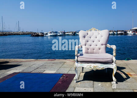 Una poltrona con decorazioni barocche in piedi sul cemento in primo piano. Una porta di fisher, barche e il mare Mediterraneo sono visti sullo sfondo, Foto Stock