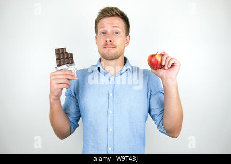 Giovane uomo bello tenendo premuto barra di cioccolato in una mano e la mela fresco in un altro isolato su sfondo bianco. Foto Stock