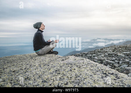 Escursionista uomo seduto in una posa yoga al picco della montagna in estate. Meditazione dopo una lunga salita su una montagna sullo sfondo di un cielo nuvoloso th Foto Stock