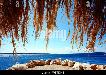 Foglie di bambù da ombrellone attraverso il quale si può vedere la spiaggia di pietra e vista della linea di orizzonte tra mare e cielo attraverso le foglie essiccate di Palm Foto Stock