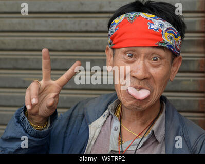 Divertente di mezza età uomo tailandese con una colorata bandana bastoni fuori la sua lingua e visualizza il segno V con la sua mano destra. Foto Stock