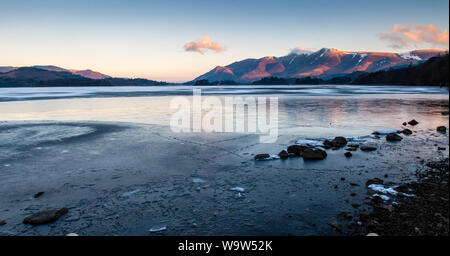Inverno alba illumina la neve Piste spolverato di Skiddaw montagna sopra un congelati Derwent Water in Inghilterra del Lake District. Foto Stock