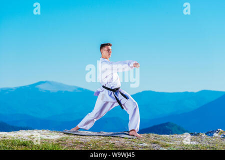 Il Karate uomo in un kimono esegue una mano anteriore kick (Choku-zuki) mentre si sta in piedi sul prato verde sulla cima di una montagna. Foto Stock