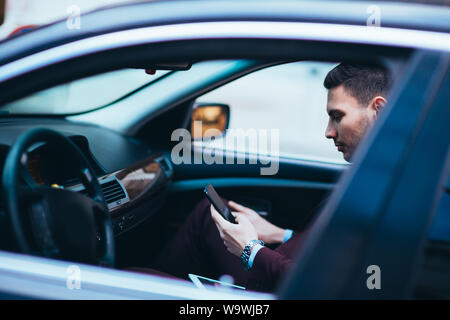 Giovane imprenditore guardando al suo cellulare stando comodamente seduti sul sedile anteriore nella sua limousine di lusso. Foto Stock