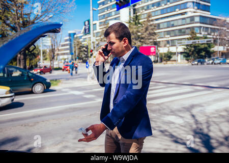 Giovane imprenditore sta parlando sul suo telefono cellulare mentre guardando sotto il cofano sollevato della sua vettura. Foto Stock