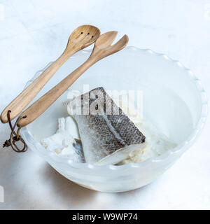 Salate e secche pesce Merluzzo bianco (Gadus morhua) in recipiente di plastica riempiti con acqua per desalinizzare e di essere cotti e cucchiaio di legno su sfondo bianco. Cibo sano. Foto Stock