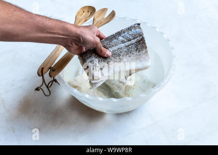 Salate e secche pesce Merluzzo bianco (Gadus morhua) in recipiente di plastica riempiti con acqua per desalinizzare e di essere cotti e cucchiaio di legno su sfondo bianco. Cibo sano. Foto Stock