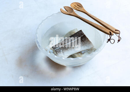 Salate e secche pesce Merluzzo bianco (Gadus morhua) in recipiente di plastica riempiti con acqua per desalinizzare e di essere cotti e cucchiaio di legno su sfondo bianco. Cibo sano. Foto Stock