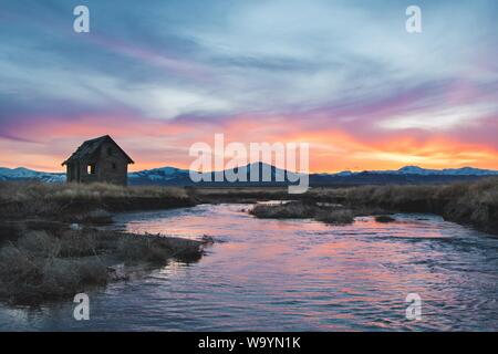 una piccola cabina sul lago circondata da piante secche e le montagne sotto il cielo durante il tramonto Foto Stock