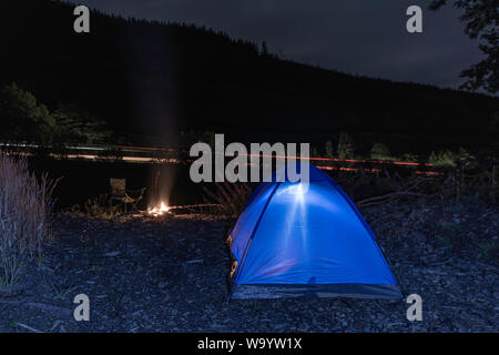 Illuminato tenda blu si accamparono nel deserto di notte.falò e sentieri di luce dalle vetture di passaggio sulla strada vicina. Campeggio selvaggio in Galles,UK.avventura. Foto Stock