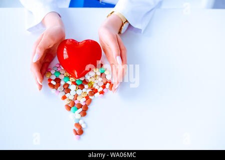 Variopinte pillole su un tavolo a forma di cuore su uno sfondo bianco. Cuore rosso . Dottore le mani sul tavolo vicino al cuore e pillole. Concetto Foto Stock