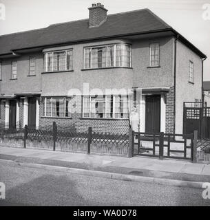 Degli anni Cinquanta, storico, lady in piedi fuori dell'entrata di 1930 costruita casa bifamiliare, con un pebbledash o copertura roughcast sul livello superiore e la parete laterale, Londra, Inghilterra, Regno Unito. Questo rivestimento di intonaco sulle pareti esterne è stata spesso applicata alle case costruite negli anni venti e trenta per motivi di design e stile e aggiunto rudimentale per la protezione dalle intemperie, ma decllined in popolarità in epoche successive. Foto Stock