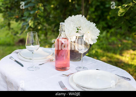 Piatti da portata con stoviglie sul panno bianco all'aperto. Tavolo in giardino con fiori e una bottiglia di bevanda. Concetto di Romantico set di tavola Foto Stock