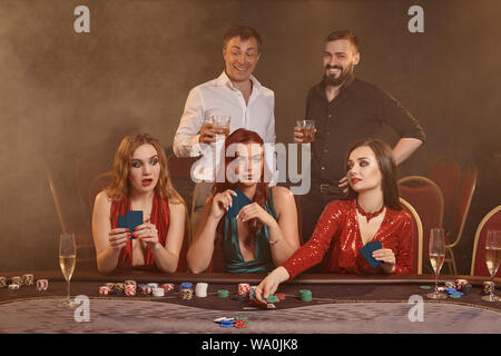 Concentrato di amici stanno giocando a poker al Casino. Golden giovani sono scommettitori in attesa di una grande vittoria in posa a tavola contro un fumo scuro ba Foto Stock