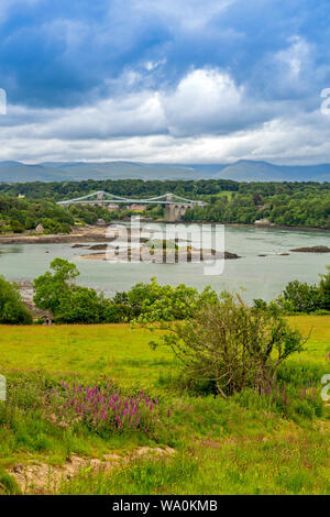 La storica 1826 Menai Bridge da Thomas Telford prende la A5 strada sopra il Menai Straits in Anglesey, con le colline di Snowdonia oltre, Wales, Regno Unito Foto Stock