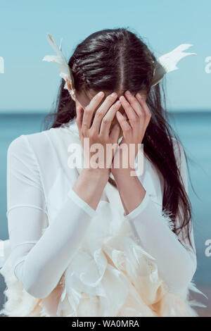 Turbare la donna a White Swan costume in piedi sul fiume azzurro cielo e sfondo, che ricopre la faccia con le mani Foto Stock