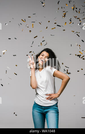 Giovane americano africano donna sorridenti vicino shiny coriandoli mentre in piedi con la mano sul hip e che mostra segni di pace su grigio Foto Stock