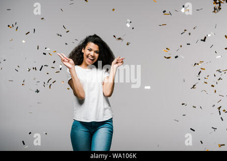 Giovane americano africano donna sorridenti vicino a cadere coriandoli mentre permanente e la mostra segno di pace su grigio Foto Stock