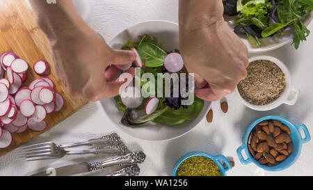 Mix di erbe con insalata fresca di ravanello organico, lattughe e spinaci, alcuni condimento, mandorle e olio vegetale. Close up su sfondo bianco, Donna mani Foto Stock