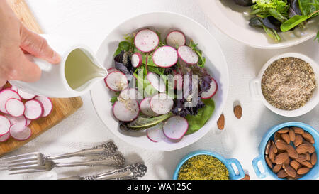 Mix di erbe con insalata fresca di ravanello organico, lattughe e spinaci, alcuni condimento, mandorle e olio vegetale. Close up su sfondo bianco, Donna mani Foto Stock