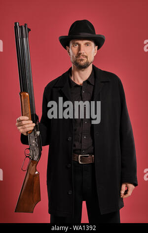 Studio shot di una bella persona con barba, baffi, indossa giacca nera e cappello, in possesso di una pistola mentre posa contro uno sfondo rosso con copia s Foto Stock