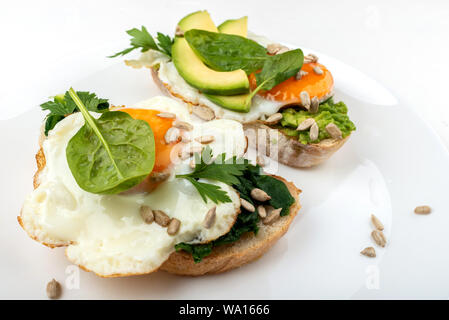 Uova fritte su un tosta pane con avocado, spinaci e semi su una piastra bianca su sfondo bianco Foto Stock