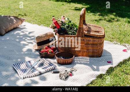 Cesto in Vimini con rose e vino su coltre bianca vicino a cappello di paglia, posate sul tovagliolo e bacche al giorno di sole in giardino Foto Stock