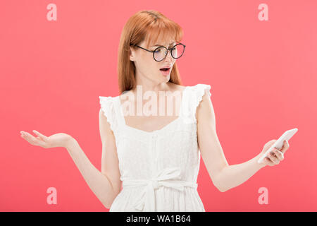 Orinato redhead donna in bicchieri e abito bianco utilizza lo smartphone isolato in rosa Foto Stock