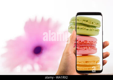 Mano che tiene il telefono e prendendo la foto del gustoso macaron colorati in piastra sul pastello alla moda con sfocato fiore rosa, spazio per il testo. Moderno cibo foto Foto Stock