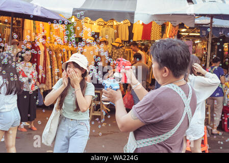 Bangkok, Thailandia: giovane donna ride nascondendo il suo viso con le mani nel cloud di bolle di sapone inviato dal venditore di soffianti di bolla al famoso mercato di Chatuchak Foto Stock