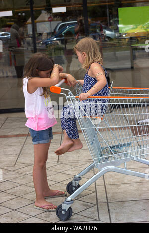 Due ragazze infront shopping mall, uno di loro è seduto nel carrello della spesa Foto Stock