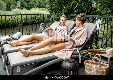 Due giovani donne in accappatoio sdraiati sui lettini rilassanti e trascorrere il tempo in una spa di lusso sulla terrazza all'aperto Foto Stock