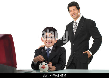 Ragazzo imitando come un uomo d'affari e mettendo la moneta in una piggybank con suo padre Foto Stock