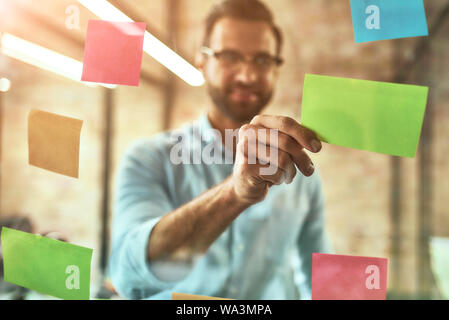 Kanban board. Giovane e bello uomo barbuto in occhiali utilizzando adesivi colorati e sorridenti mentre in piedi nella parte anteriore della parete di vetro. Gestione del progetto Foto Stock