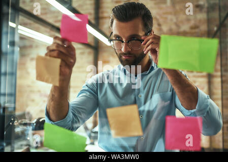 Idee fresche. Giovane e bello imprenditore in occhiali di pianificazione del processo di lavoro e utilizzo di adesivi colorati mentre in piedi nella parte anteriore della parete di vetro. Gestione del progetto Foto Stock