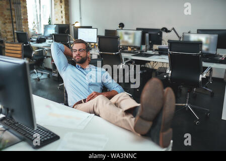 Il tempo per rilassarsi. Bello barbuto giovane uomo in occhiali e usura formale tenendo la mano dietro la sua testa e mantenendo le gambe sul tavolo. Ufficio moderno. Il concetto di business Foto Stock