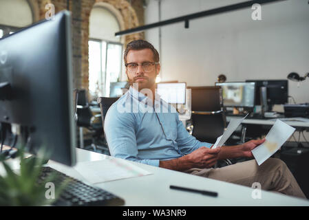 Controllare il report. Occupato uomo barbuto in occhiali e abbigliamento formale dei documenti di contenimento e guardando il monitor di un computer mentre è seduto in un ufficio moderno. Il concetto di business. Concetto di lavoro Foto Stock