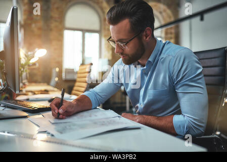 Giovane ingegnere. Occupato di successo uomo barbuto in occhiali e formale del disegno di usura qualcosa mentre si lavora in ufficio. Ingegnere. Piano di costruzione. Progetto architettonico Foto Stock