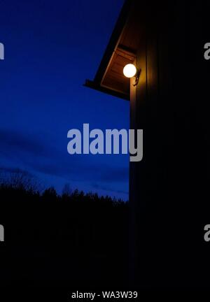 colpo di una lampadina illuminata su una cabina di legno vicino alberi sotto un cielo blu chiaro durante la notte Foto Stock