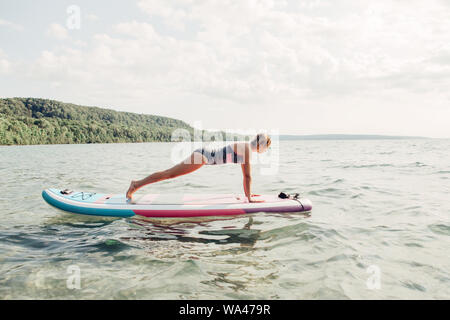 Medioevo donna caucasica la pratica dello yoga sulla paletta sup tavola da surf al tramonto. Femmina facendo stretching allenamento sul lago di acqua. Moderno tanga individuali Foto Stock